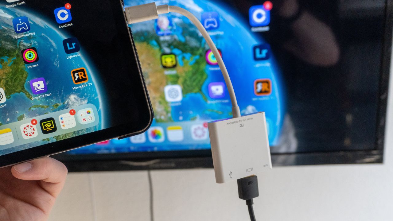 A white iPad HDMI adaptor is plugged into an iPad. An HDMI cable hangs from the adaptor and the screen is mirroring the display on the TV. 